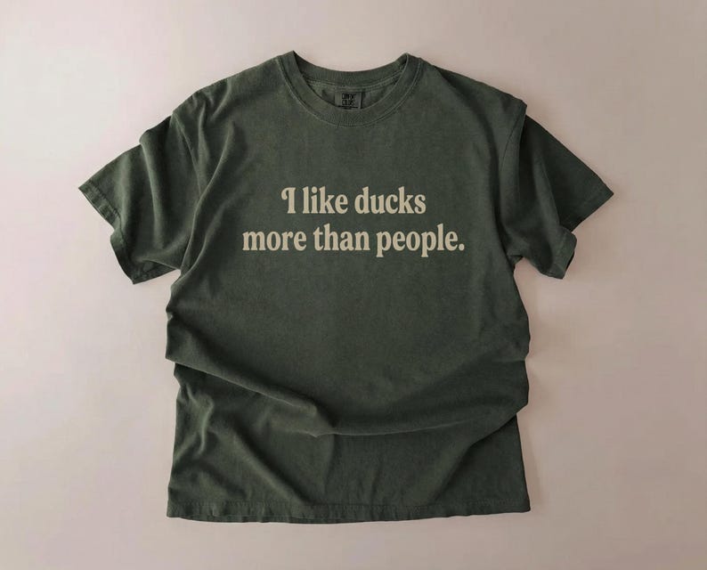 May include: Olive green t-shirt with the text "I like ducks more than people" in a white, sans-serif font. The shirt has short sleeves and a crew neck. The fabric appears to be soft and slightly faded.