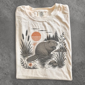 Könnte beinhalten: Ein cremefarbenes T-Shirt mit einem Grafikdruck eines Bibers in einem Teich mit Rohrkolben, Kiefern und einer orangefarbenen Sonne. Das Design enthält eine kleine orangefarbene Blume im Vordergrund. Das T-Shirt hat ein Comfort Colors Etikett.
