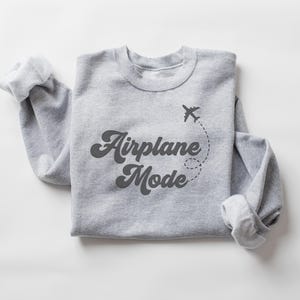 K&ouml;nnte beinhalten: Ein hellgraues Sweatshirt mit dem Schriftzug "Airplane Mode" in dunkler, retro Schrift. Eine kleine Flugzeug-Grafik mit gepunkteter Flugbahn befindet sich &uuml;ber dem Text. Die &Auml;rmel sind umgeschlagen.