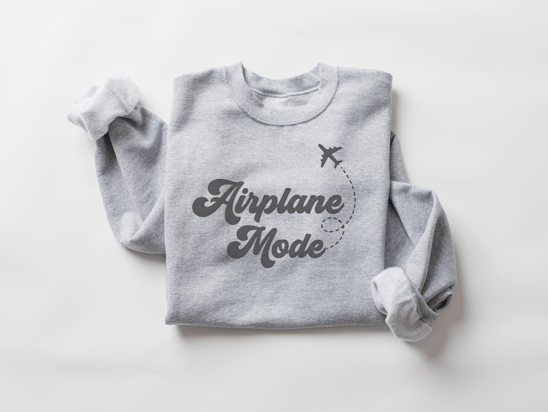 K&ouml;nnte beinhalten: Ein hellgraues Sweatshirt mit dem Schriftzug "Airplane Mode" in dunkler, retro Schrift. Eine kleine Flugzeug-Grafik mit gepunkteter Flugbahn befindet sich &uuml;ber dem Text. Die &Auml;rmel sind umgeschlagen.