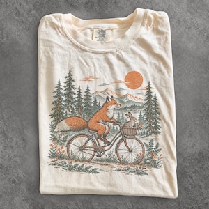Peut inclure: Un t-shirt couleur crème avec une impression graphique représentant un renard à vélo transportant un lapin dans le panier avant. Le décor inclut des pins, des montagnes et un soleil orange. L'étiquette Comfort Colors est visible au col.