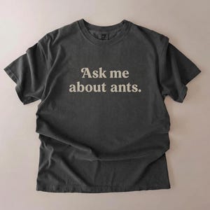 Op de afbeelding: Donkergrijs t-shirt met de tekst "Ask me about ants" in een vintage lettertype. Het shirt heeft een ronde hals en korte mouwen. Het kledingstuk is gemaakt van zacht materiaal en ligt op een neutrale achtergrond.