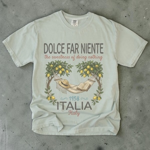 Puede incluir: Una camiseta azul claro con un dise&ntilde;o gr&aacute;fico que presenta una hamaca, un sombrero y un libro bajo limoneros. El texto dice "DOLCE FAR NIENTE the sweetness of doing nothing" y "1958 ITALIA Italy".