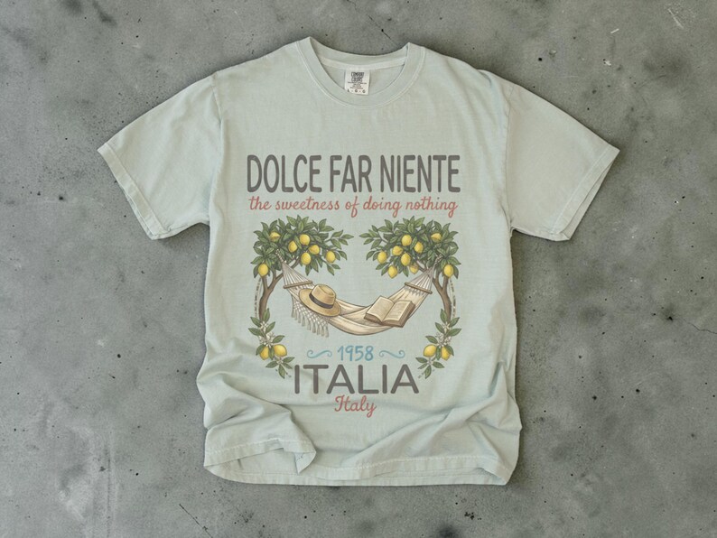 Puede incluir: Una camiseta azul claro con un dise&ntilde;o gr&aacute;fico que presenta una hamaca, un sombrero y un libro bajo limoneros. El texto dice "DOLCE FAR NIENTE the sweetness of doing nothing" y "1958 ITALIA Italy".