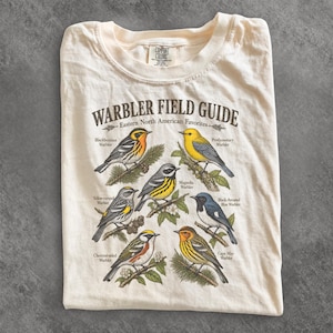 May include: A cream-coloured t-shirt featuring a Warbler Field Guide graphic. The print displays six Eastern North American warblers perched on branches, labelled with their names: Blackburnian, Prothonotary, Magnolia, Yellow-rumped, Black-throated Blue, and Cape May.