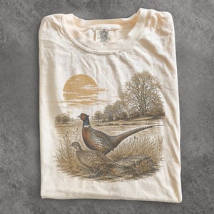 Puede incluir: Una camiseta de color crema con un estampado gráfico de dos faisanes en un campo de hierba con árboles y un gran sol de fondo. La camiseta tiene cuello redondo y una etiqueta de la marca Comfort Colors impresa en el interior del cuello.