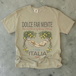 Puede incluir: Camiseta color beige con la frase "DOLCE FAR NIENTE" y "the sweetness of doing nothing". El dise&ntilde;o incluye una hamaca, un sombrero y un libro bajo limoneros, con "ITALIA" y el a&ntilde;o "1958" impresos debajo. La camiseta es de color neutro.