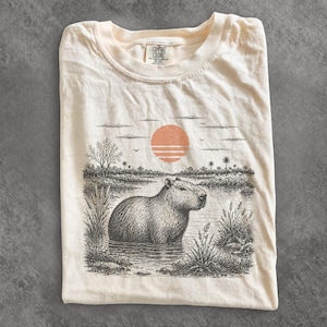 Puede incluir: Una camiseta de color crema con una ilustración en tinta negra y blanca de un capibara en el agua. Sobre el animal hay un sol de color melocotón con líneas horizontales. La camiseta tiene una etiqueta de la marca Comfort Colors en el cuello.