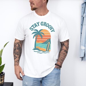 May include: White t-shirt with a tropical graphic. The design features a palm tree, surfboard, and ocean scene with the words "Stay Groovy" in teal. The shirt is made of a soft material.
