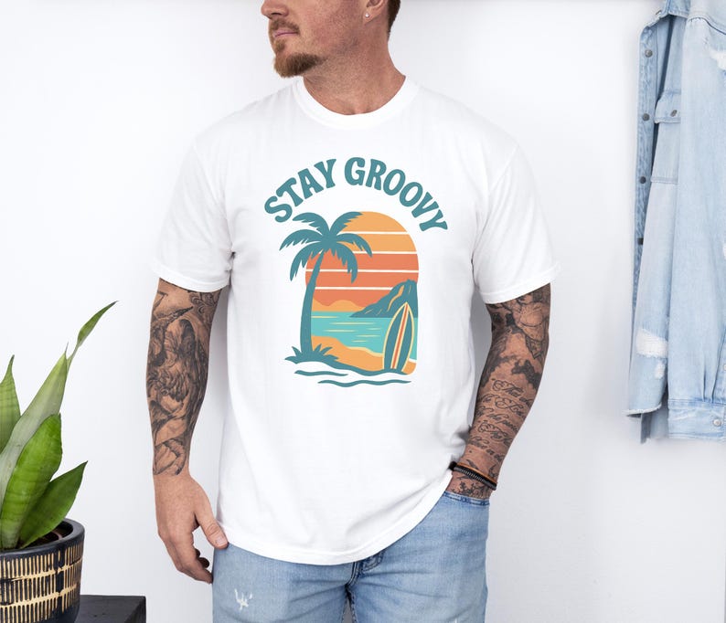 May include: White t-shirt with a tropical graphic. The design features a palm tree, surfboard, and ocean scene with the words "Stay Groovy" in teal. The shirt is made of a soft material.