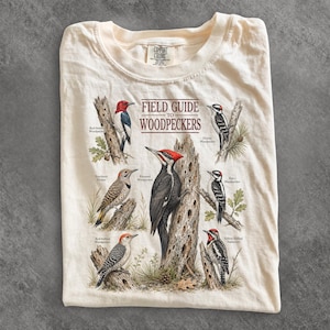 May include: A cream-colored t-shirt featuring a graphic print titled FIELD GUIDE TO WOODPECKERS. The design displays six detailed illustrations of various woodpecker species perched on tree trunks, each labeled with its common name.