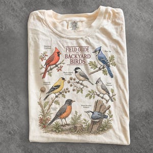 Könnte beinhalten: Ein cremefarbenes T-Shirt mit einer Grafik mit der Aufschrift Field Guide to Backyard Birds. Der Druck zeigt Illustrationen eines Kardinals, eines Blauhähers, eines Goldzeisigs, einer Schwarzkopfmeise, einer Tufted-Meise, einer Wanderdrossel und eines Weißbrustkleibers.