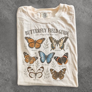 Puede incluir: Una camiseta de color crema con un gráfico titulado BUTTERFLY FIELD GUIDE Wings of the Wild. El estampado muestra seis ilustraciones de mariposas etiquetadas: Monarch, Swallowtail, Painted Lady, Blue Morpho, Red Admiral, Glasswing y Common Buckeye.