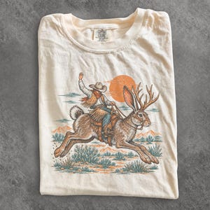 May include: A cream-coloured short-sleeve t-shirt featuring a graphic print of a cowboy riding a jackalope. The illustration depicts a desert landscape with mountains, cacti, and an orange sun. The shirt has a Comfort Colors brand label at the neck.