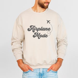K&ouml;nnte beinhalten: Beiges Sweatshirt mit dem Text "Airplane Mode" in Retro-Schrift, begleitet von einer kleinen Flugzeuggrafik und gepunkteter Flugbahn. Das lang&auml;rmlige Oberteil mit Rundhalsausschnitt ist neutral gef&auml;rbt.