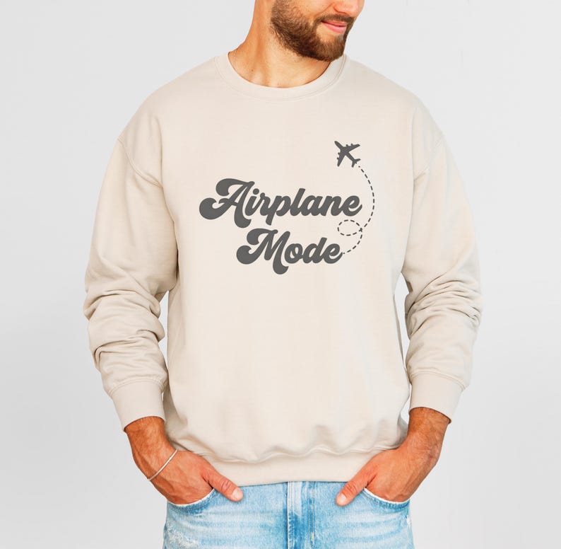 K&ouml;nnte beinhalten: Beiges Sweatshirt mit dem Text "Airplane Mode" in Retro-Schrift, begleitet von einer kleinen Flugzeuggrafik und gepunkteter Flugbahn. Das lang&auml;rmlige Oberteil mit Rundhalsausschnitt ist neutral gef&auml;rbt.