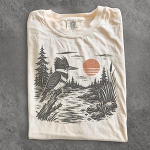 以下が含まれることがあります： クリーム色のTシャツで、川辺の枝にとまるカワセミ、松の木、ガマ、大きなオレンジ色の太陽が描かれた白黒のグラフィックプリントが施されています。首元の内側のラベルにはComfort Colorsの文字があります。