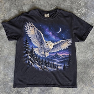 Könnte beinhalten: Ein schwarzes Kurzarm-T-Shirt mit einem Grafikdruck einer weißen Schnee-Eule im Flug über einer dunklen Wald- und Berglandschaft unter einem Sternenhimmel mit einer Mondsichel. Das Motiv befindet sich mittig auf der Brustpartie.