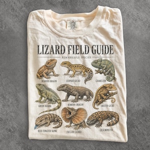 Puede incluir: Una camiseta de color crema con un gráfico titulado LIZARD FIELD GUIDE REMARKABLE SPECIES. El estampado muestra ilustraciones de un dragón barbudo, gecko leopardo, camaleón, iguana verde, dragón de Komodo, gecko crestado, eslizón de lengua azul, lagarto de gorguera y monstruo de Gila con sus nombres impresos debajo.