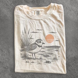 Może przedstawiać: Kremowy t-shirt z nadrukiem graficznym przedstawiającym ptaka brzegowego stojącego na plaży z wysoką trawą i zachodzącym słońcem. Etykieta marki Comfort Colors jest widoczna przy wewnętrznym dekolcie. Ilustracja wykonana w kolorze ciemnoszarym i brzoskwiniowym.
