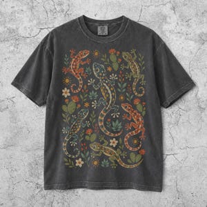 以下が含まれることがあります： チャコールグレーの半袖Tシャツ。中央には5匹のカラフルなトカゲと、サボテンや小さな花などの様々な砂漠の植物が描かれたグラフィックプリントが施されており、前面全体に繰り返し模様として配置されています。