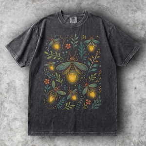 Puede incluir: Una camiseta de manga corta gris carbón con un estampado gráfico de varias luciérnagas con abdómenes amarillos brillantes, rodeadas de delicado follaje verde y pequeñas flores naranjas. La camiseta tiene cuello redondo y un acabado desgastado.