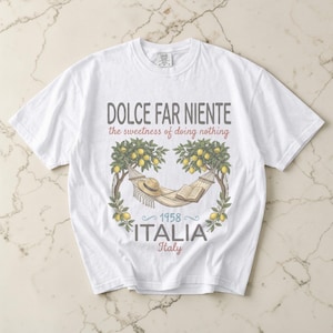 Puede incluir: Camiseta blanca con un gr&aacute;fico que presenta una hamaca, un sombrero y un libro entre limoneros. El texto incluye "DOLCE FAR NIENTE" y "ITALIA 1958 Italy". El dise&ntilde;o tiene una est&eacute;tica vintage con tem&aacute;tica de viajes.