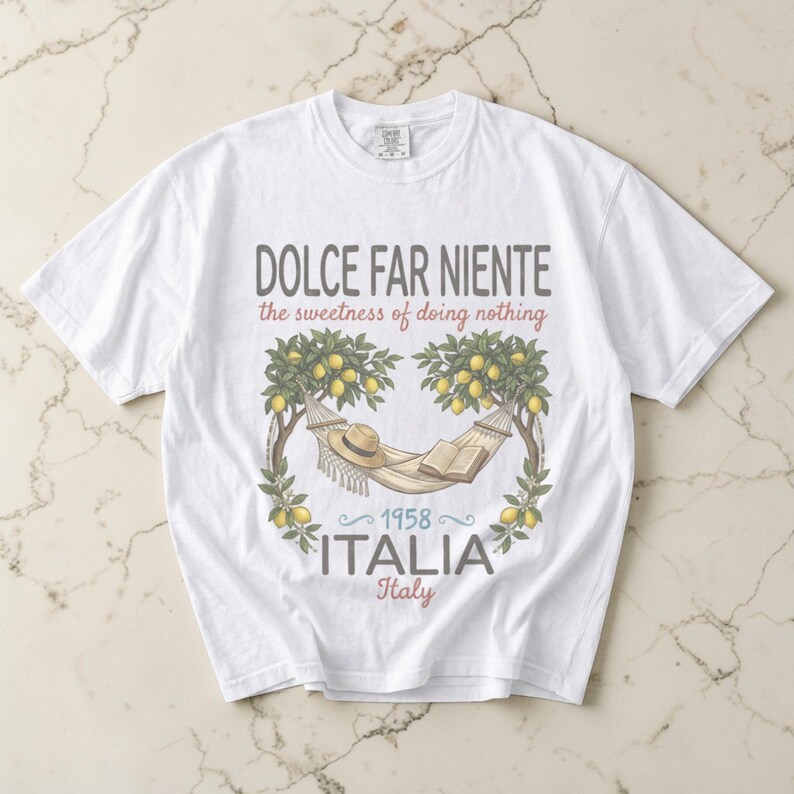 Puede incluir: Camiseta blanca con un gr&aacute;fico que presenta una hamaca, un sombrero y un libro entre limoneros. El texto incluye "DOLCE FAR NIENTE" y "ITALIA 1958 Italy". El dise&ntilde;o tiene una est&eacute;tica vintage con tem&aacute;tica de viajes.