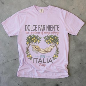 Puede incluir: Camiseta lila claro con las palabras "DOLCE FAR NIENTE" y "the sweetness of doing nothing". El dise&ntilde;o incluye una hamaca, un sombrero y un libro bajo limoneros, con "1958 ITALIA Italy" debajo.