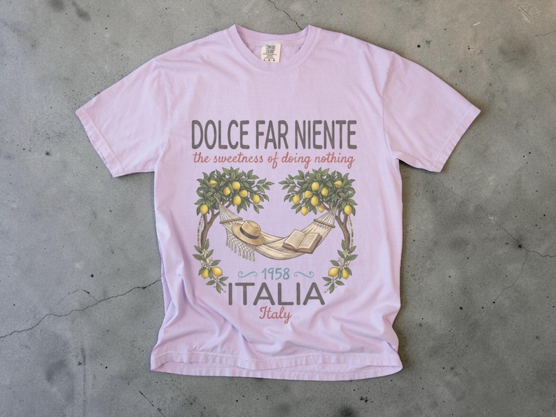 Puede incluir: Camiseta lila claro con las palabras "DOLCE FAR NIENTE" y "the sweetness of doing nothing". El dise&ntilde;o incluye una hamaca, un sombrero y un libro bajo limoneros, con "1958 ITALIA Italy" debajo.