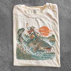 Op de afbeelding: Een crèmekleurig t-shirt met een grafische illustratie van een persoon met een cowboyhoed en chaps die op een grote meerval in een rivier rijdt. De scène bevat een zonsondergang, lisdodden en opspattend water in de rivier.