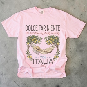 Puede incluir: Camiseta rosa claro con un dise&ntilde;o gr&aacute;fico. El dise&ntilde;o incluye una hamaca, un sombrero y un libro bajo limoneros. El texto dice "DOLCE FAR NIENTE the sweetness of doing nothing" y "1958 ITALIA Italy".