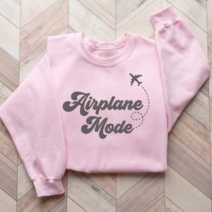 K&ouml;nnte beinhalten: Hellrosa Sweatshirt mit dem Schriftzug "Airplane Mode" in dunkelgrauer Schrift. Ein kleines Flugzeug-Symbol mit gestrichelter Flugbahn erg&auml;nzt das Design. Das gefaltete Sweatshirt liegt auf einer hellbraunen Holzoberfl&auml;che.