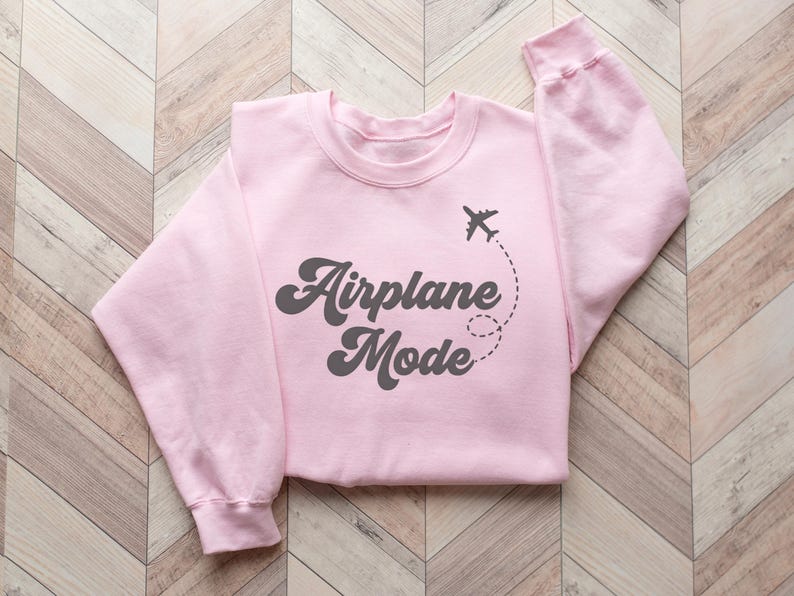 K&ouml;nnte beinhalten: Hellrosa Sweatshirt mit dem Schriftzug "Airplane Mode" in dunkelgrauer Schrift. Ein kleines Flugzeug-Symbol mit gestrichelter Flugbahn erg&auml;nzt das Design. Das gefaltete Sweatshirt liegt auf einer hellbraunen Holzoberfl&auml;che.