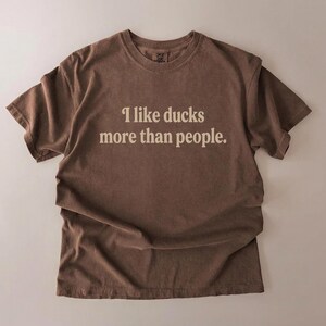 May include: A brown t-shirt with the text "I like ducks more than people" in a white, sans-serif font. The shirt is made of a soft material and has a relaxed fit.