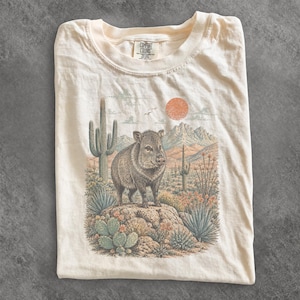 Może przedstawiać: Kremowy t-shirt z grafiką przedstawiającą pekari stojące na skale w pustynnym krajobrazie. Ilustracja zawiera kaktusy saguaro, pustynną roślinność, góry i słońce. Na metce przy kołnierzu znajduje się napis Comfort Colors.