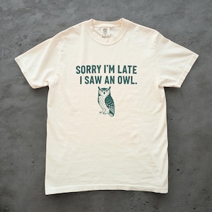 May include: Off-white t-shirt featuring the text "SORRY I'M LATE I SAW AN OWL." above a teal owl graphic. The tee has a round neck and short sleeves. The design is printed centrally on the front of the garment.