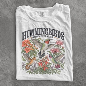 May include: A white t-shirt featuring a graphic print titled HUMMINGBIRDS GARDEN FIELD GUIDE. The design displays three hummingbirds in flight surrounded by colourful flowers and plants, with labels identifying them as Ruby-throated, Rufous, and Broad-tailed.