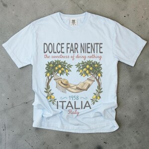 Puede incluir: Camiseta azul claro con el texto "DOLCE FAR NIENTE" y "the sweetness of doing nothing". La camiseta presenta un gr&aacute;fico de una hamaca, sombrero y libro bajo limoneros, con "1958 ITALIA Italy" debajo.