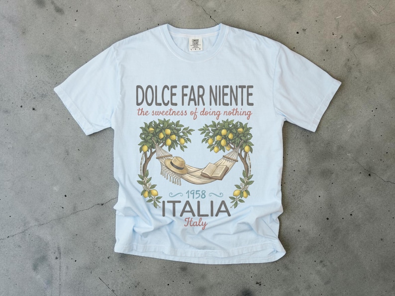 Puede incluir: Camiseta azul claro con el texto "DOLCE FAR NIENTE" y "the sweetness of doing nothing". La camiseta presenta un gr&aacute;fico de una hamaca, sombrero y libro bajo limoneros, con "1958 ITALIA Italy" debajo.