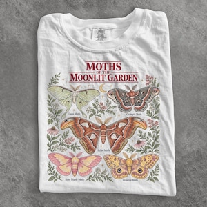Puede incluir: Una camiseta blanca con un estampado gráfico titulado MOTHS OF THE MOONLIT GARDEN. El diseño muestra cinco ilustraciones detalladas de polillas etiquetadas como Luna Moth, Cecropia Moth, Atlas Moth, Rosy Maple Moth e Imperial Moth, rodeadas de follaje botánico.