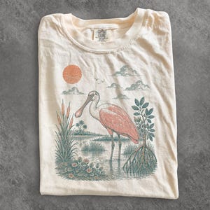 May include: A cream-colored t-shirt featuring a graphic print of a pink roseate spoonbill standing in a marshy landscape with cattails, a mangrove tree, and a textured orange sun. The inner neck label displays the text Comfort Colors.