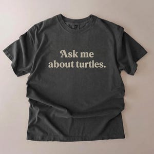 Może przedstawiać: Ciemnoszara koszulka z napisem "Ask me about turtles" w białym kolorze. Koszulka ma luźny krój i lekko wyblakły wygląd. Odzież jest ułożona płasko na neutralnym tle.