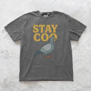 May include: Dark gray t-shirt with a graphic of a pigeon and the words "STAY COO" in yellow. The pigeon is light blue and gray with orange feet and a yellow beak. The shirt is a casual style.