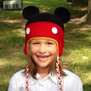 May include: A child wearing a red and black crocheted hat with Mickey Mouse ears and white buttons. The hat has long yellow, red, and white braided ties.