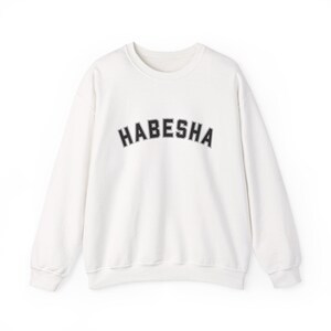 May include: A white crewneck sweatshirt with the word "HABESHA" arched across the chest in black block letters. The sweatshirt has long sleeves and a classic, casual design, suitable for everyday wear.