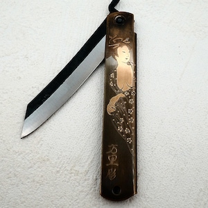 Può includere: Un coltello pieghevole color bronzo con una lama nera. Il manico presenta un disegno inciso di una donna in abiti tradizionali. Caratteri giapponesi incisi sul manico. Il coltello è aperto, rivelando la lama affilata.