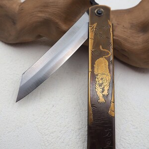 May include: A traditional Japanese folding knife with a tiger and bamboo design on the handle. The blade is made of steel and the handle is made of brass. The knife is closed and the handle has a gold inscription.