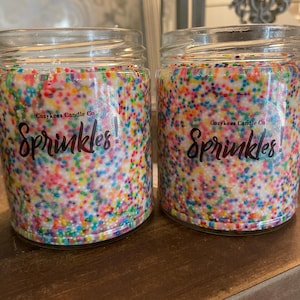 Könnte beinhalten: Zwei Glasbehälter gefüllt mit einer weißen Kerze und bunten Streuseln. Die Behälter tragen ein Etikett mit der Aufschrift "Cozykees Candle Co. Sprinkles!"