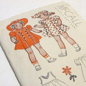 May include: Vintage McCall's sewing pattern for a child's dress. The pattern includes two different styles of dresses, one with a floral print and one with a solid orange color. The pattern is labeled "Sample" and includes instructions for making the dress.